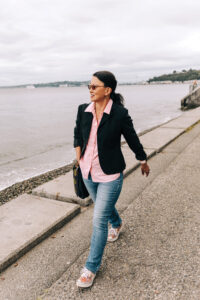 A woman in a black sweater, pink shirt, and jeans walks down a cement pathway next to a body of water.