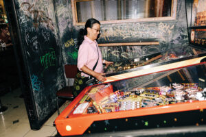 A woman in a pink shirt stands in front of a pinball machine in a room with black walls with graffiti writing.
