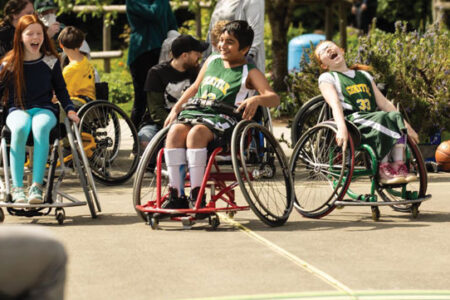 Four people in colorful clothing using wheelchairs on a paved court in the foreground with a crowd of people sitting on a bench in the background.