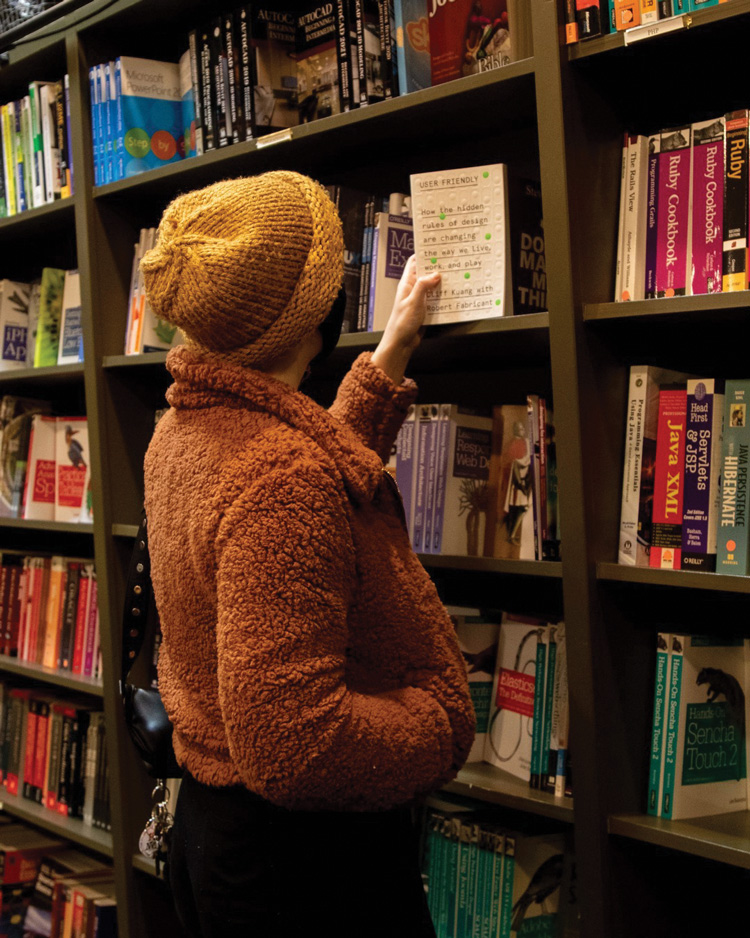 A person wearing an orange jacket and yellow beanie pulls out a book with a white cover from a bookshelf filled with books.
