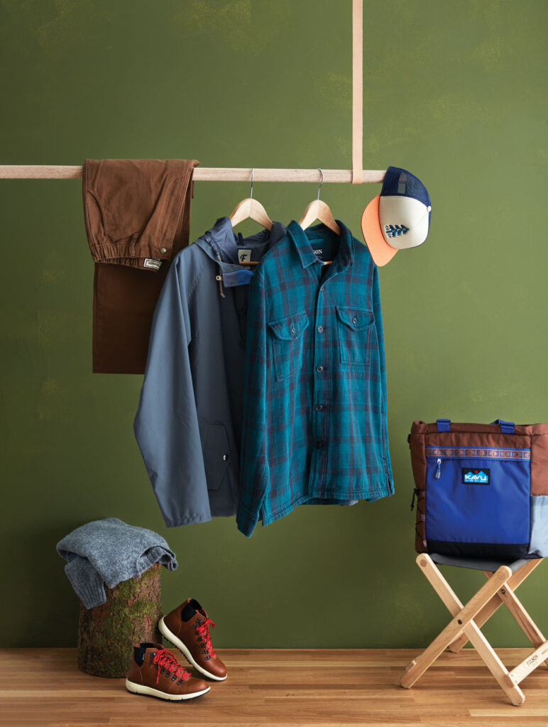 Two long-sleeve blue shirts hang next to folded brown pants and trucker hat on a clothes rack above a folded grey sweater, brown hiking boots, and a tote bag.
