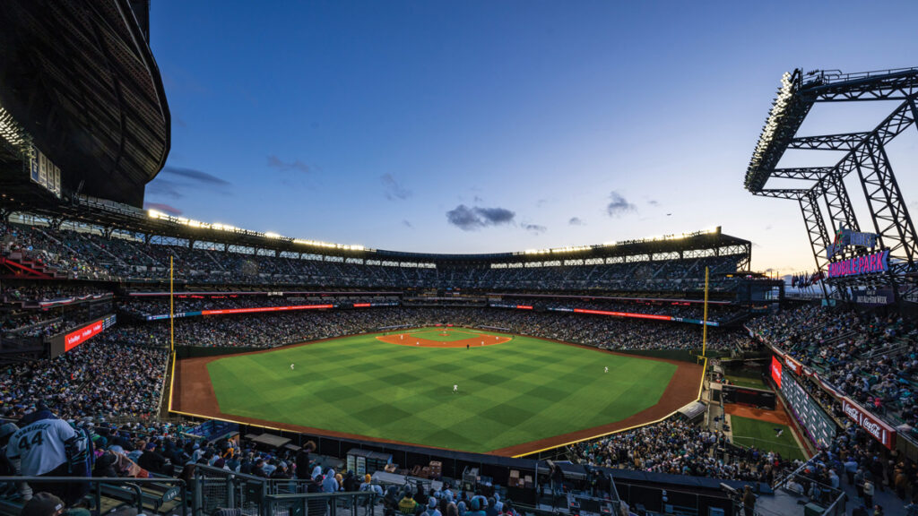 A baseball stadium at dusk showing a green grass outfield with a dirt  infield. Fans fill the seats of the stadium.