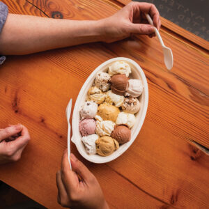 A bowl hold 14 scoops of ice cream in brown, white and pink colors. Two hands holding spoons hover over the table.