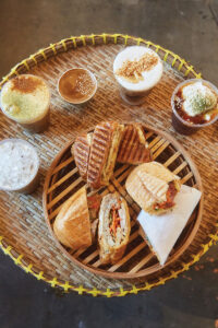 Five coffee beverages and five sandwiches sit on a woven tabletop.