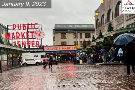 The giant Public Market Center sign is lit up against a cloudy sky, while visitors with umbrellas and rain coats cross the wet brick streets in front of the market.